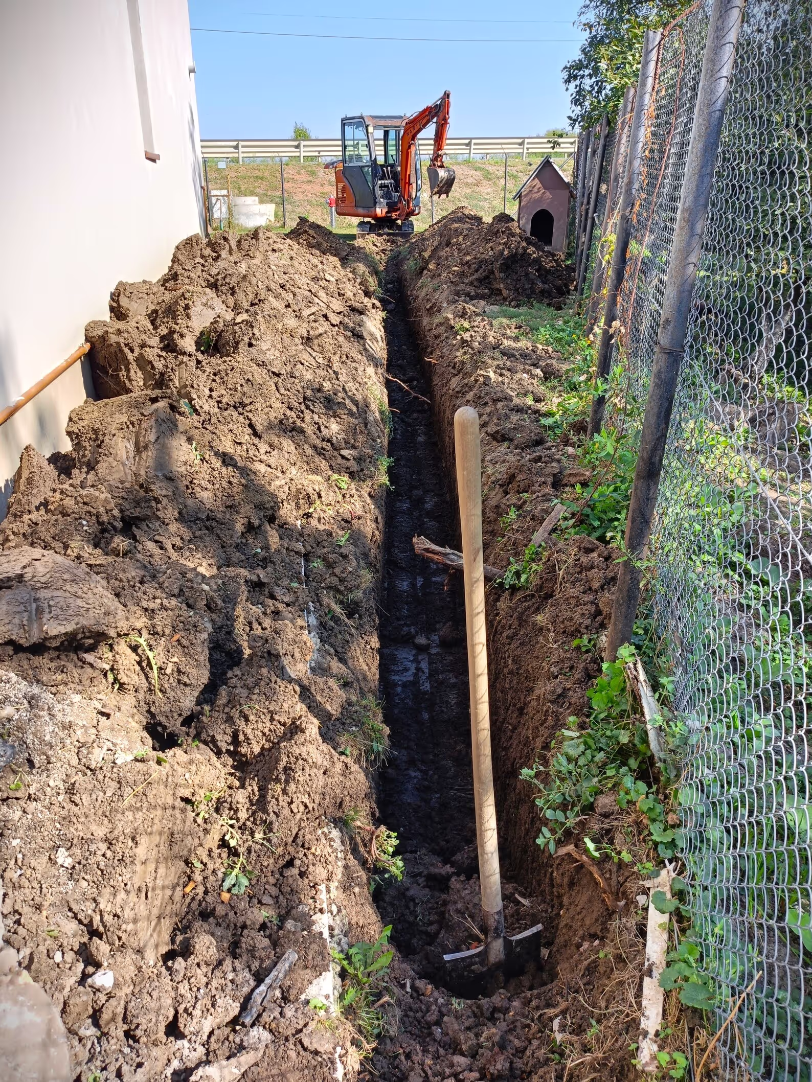 A deep trench being dug along the side of a building with an excavator at the far end, and a shovel standing upright in the trench.