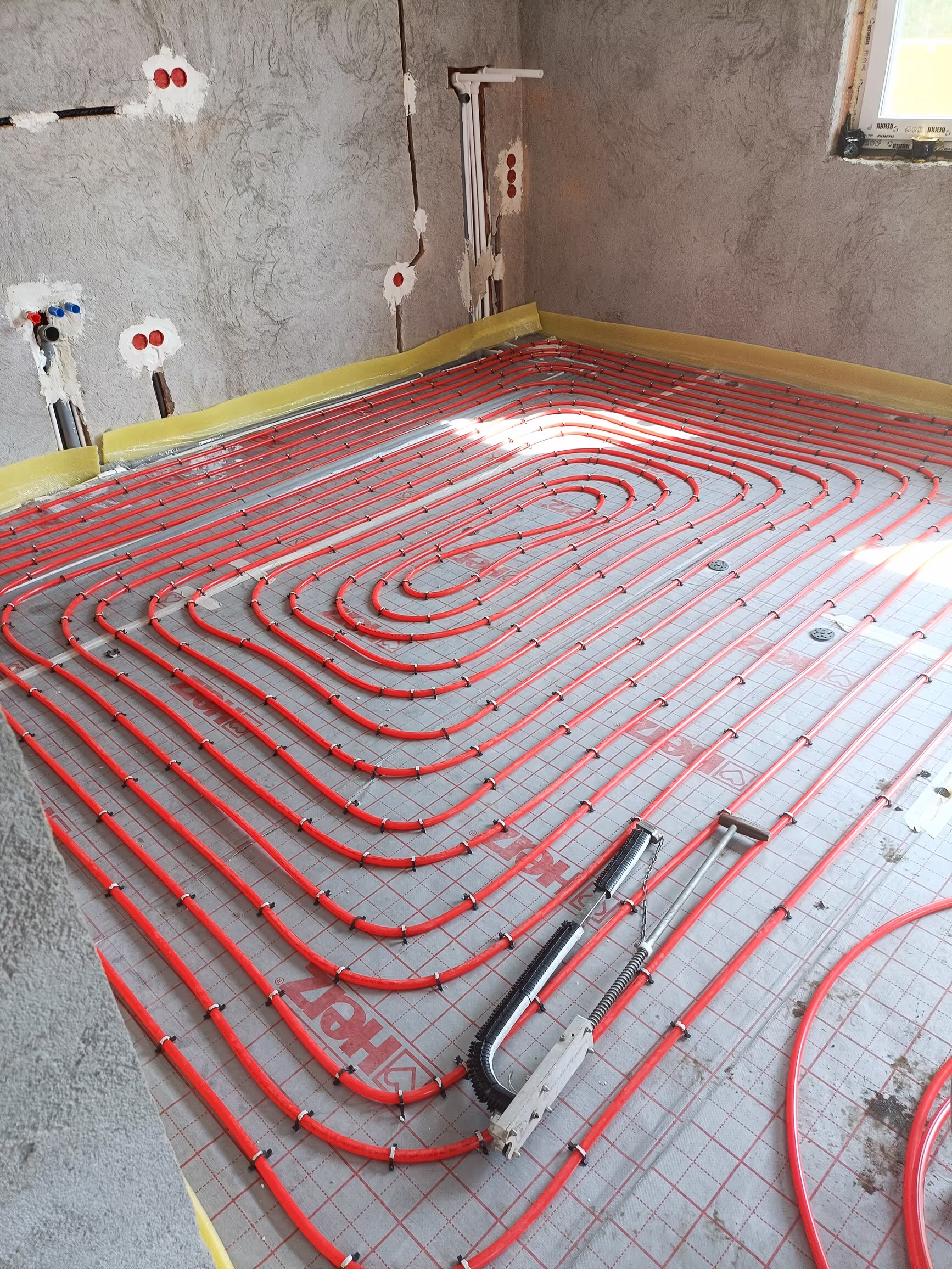 A room with underfloor heating pipes installed in a spiral pattern, red pipes secured to an insulating layer, and unfinished walls with exposed electrical outlets.
