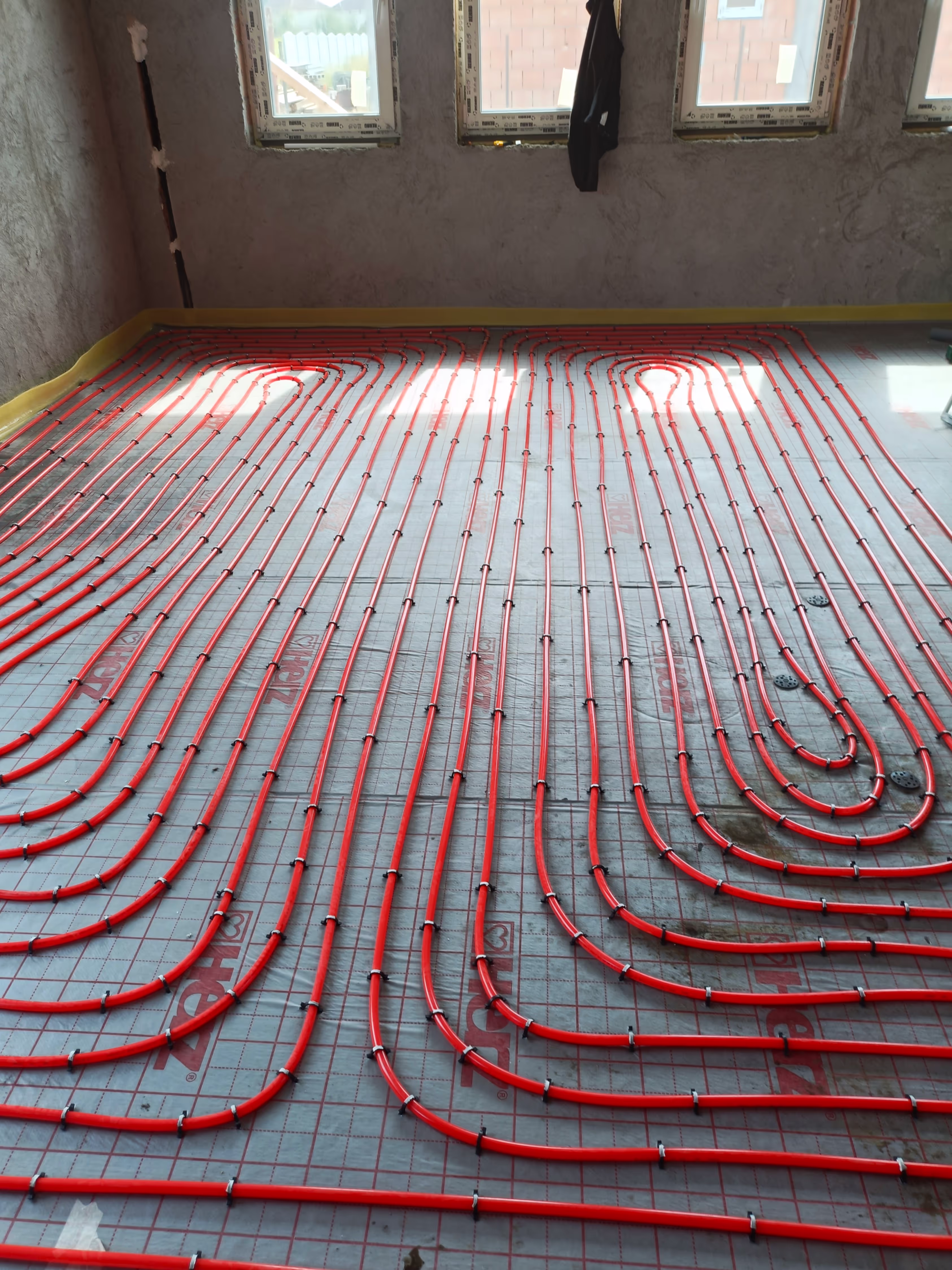 Another perspective of an underfloor heating installation, showing red pipes laid across the floor in curved patterns, with windows in the background.
