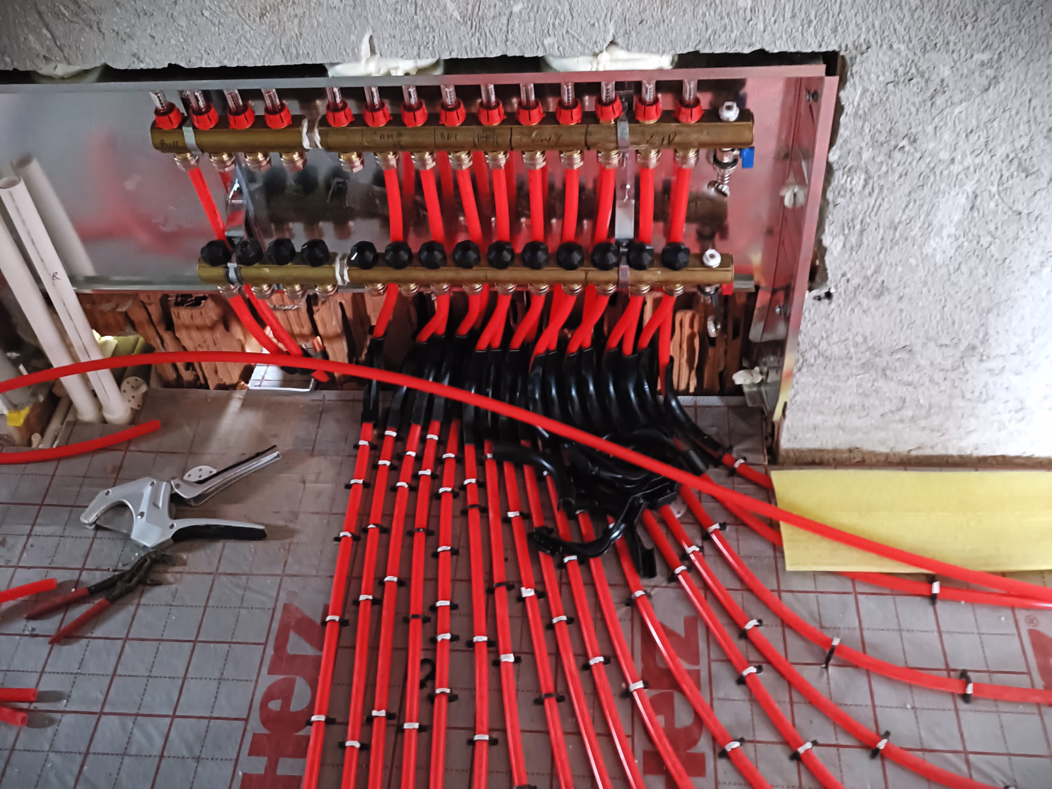 A close-up of a manifold system for underfloor heating, featuring multiple red pipes connected to a distribution panel with black and brass fittings.