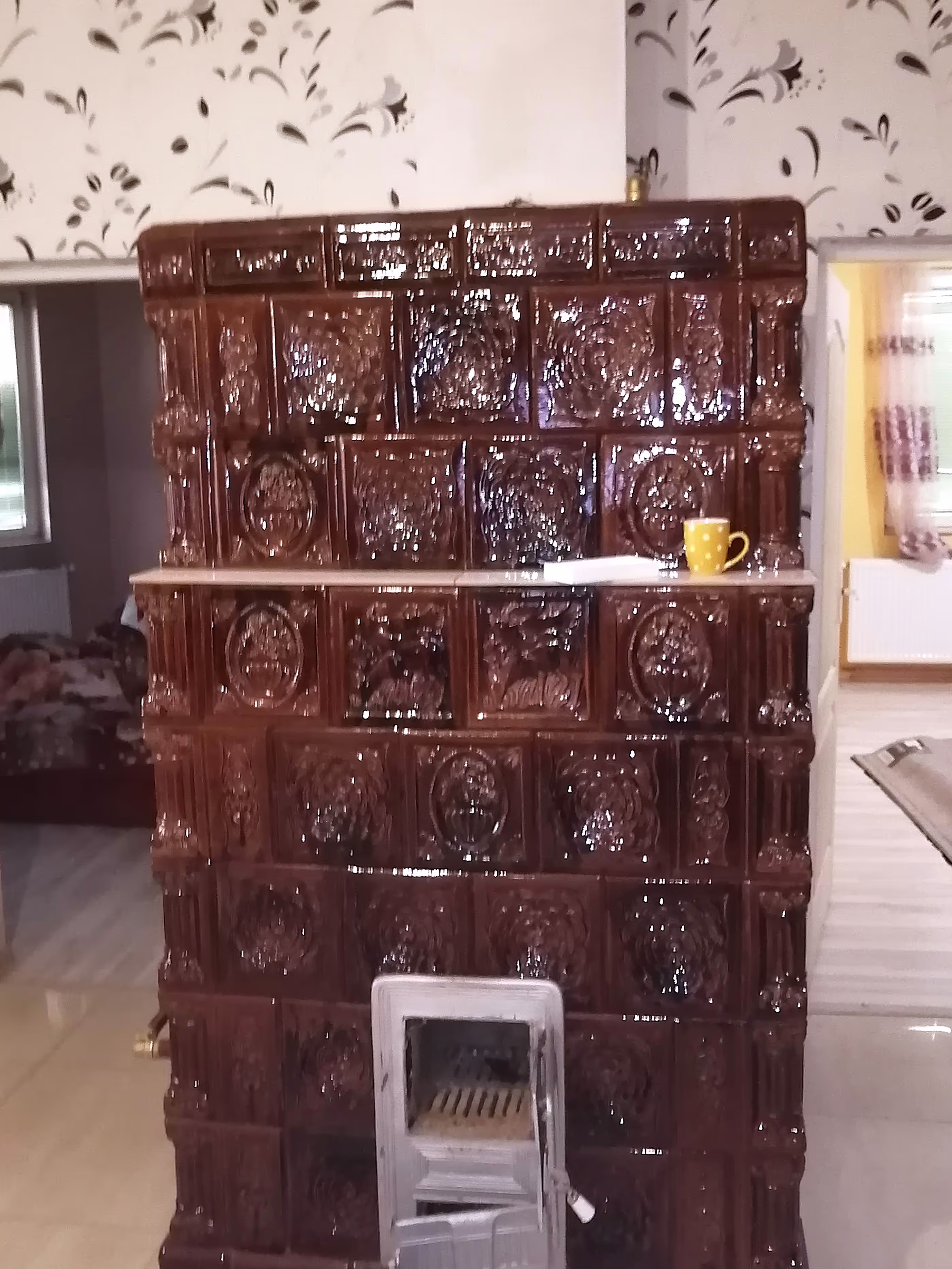 A detailed view of a glossy brown ceramic stove with an open firebox, placed in a decorated interior.