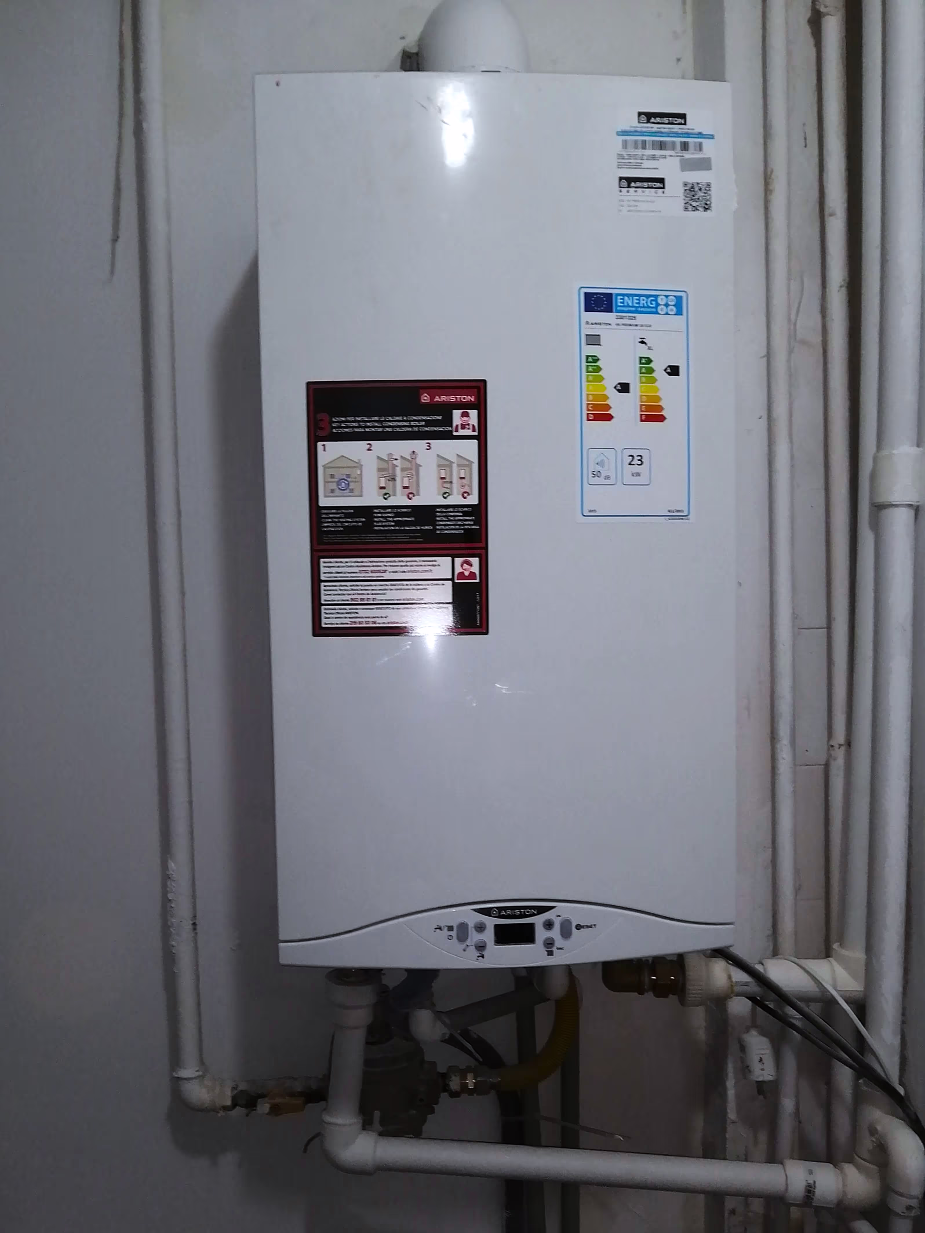 A white Ariston gas boiler mounted on a wall, with an energy efficiency label and instruction sticker. Various PVC and metal pipes are connected at the bottom.