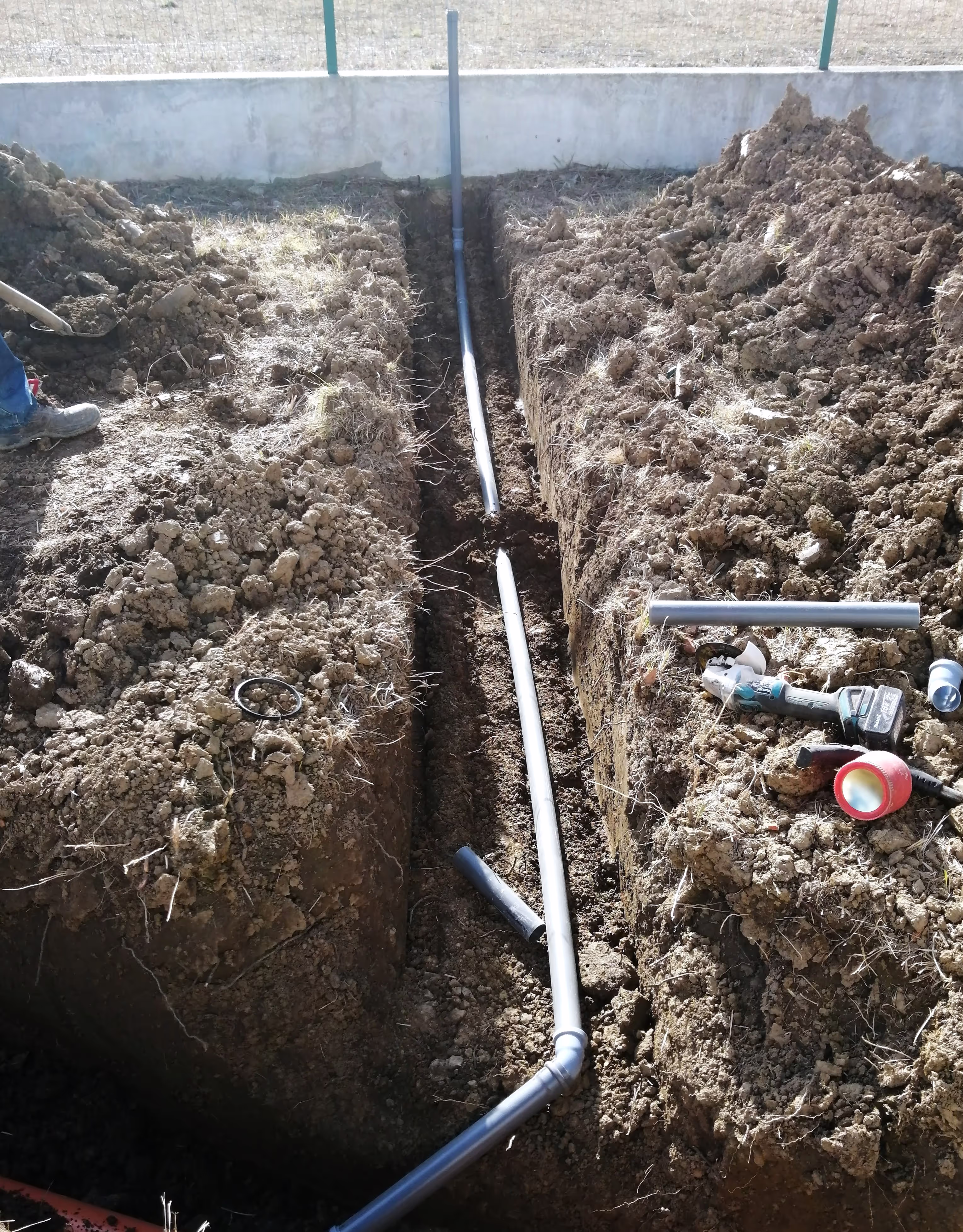 A trench with a black PVC pipe connected in sections, running along the ground. Tools and pipe fittings are scattered nearby.