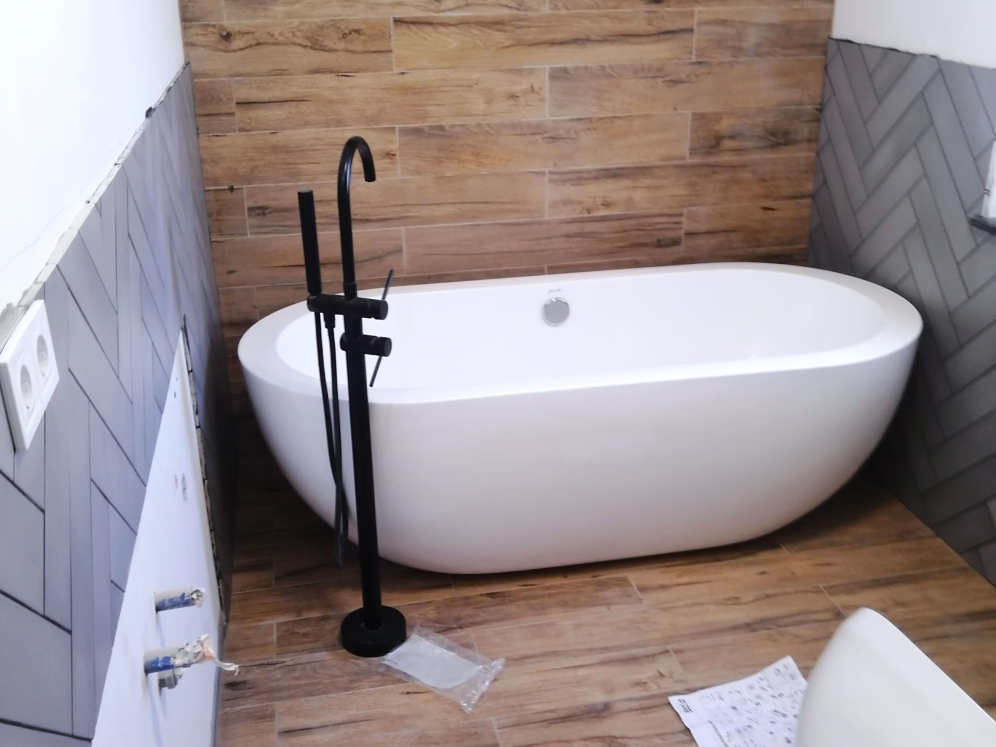 A sleek white freestanding bathtub installed in a bathroom with wooden and gray herringbone tiles, featuring a black floor-mounted faucet.