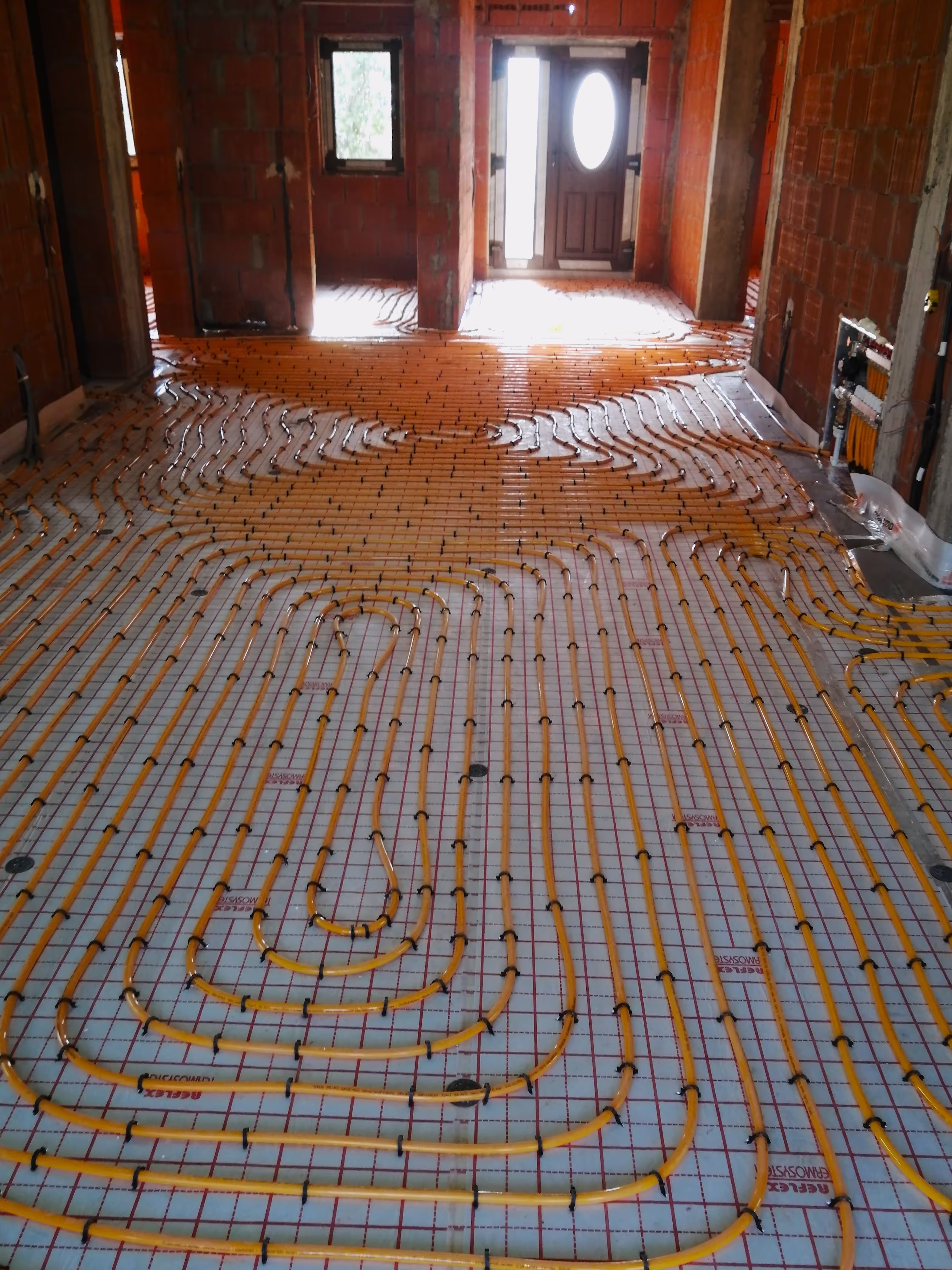A spacious room under construction with extensive underfloor heating pipes arranged in a structured pattern across the floor.