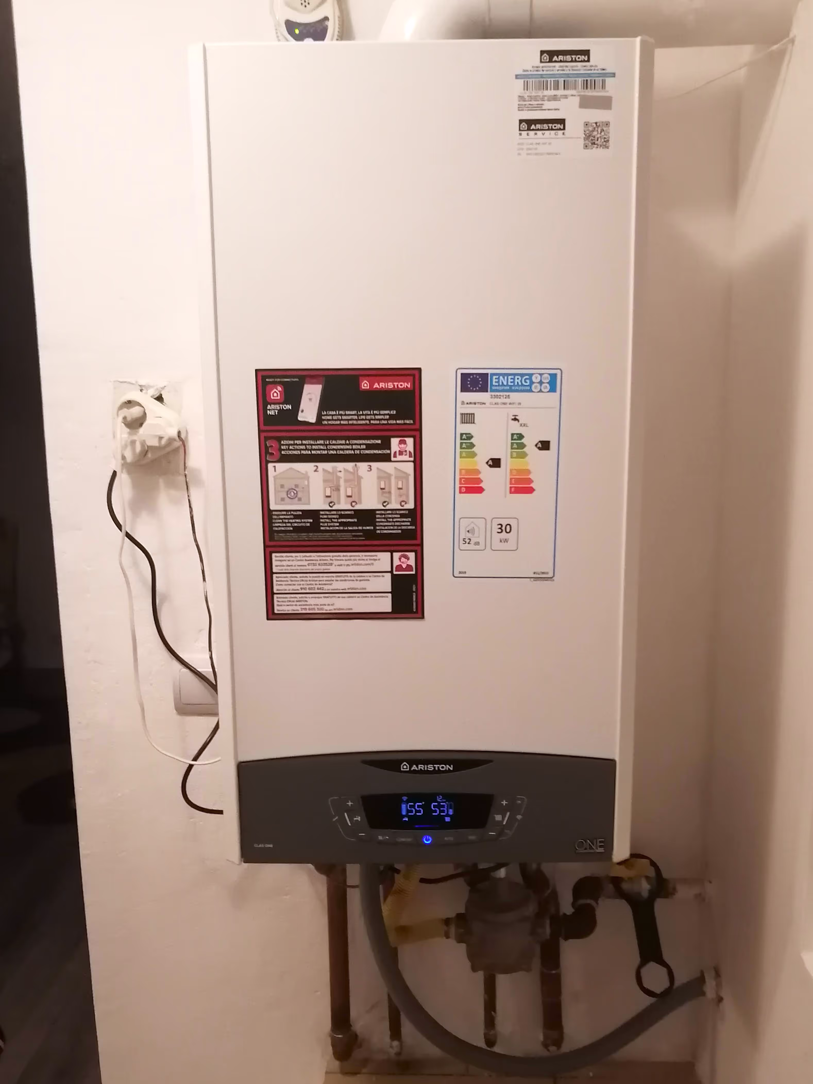 A white Ariston wall-mounted gas boiler with energy efficiency labels, installed in a utility area with visible pipes connected to the system.