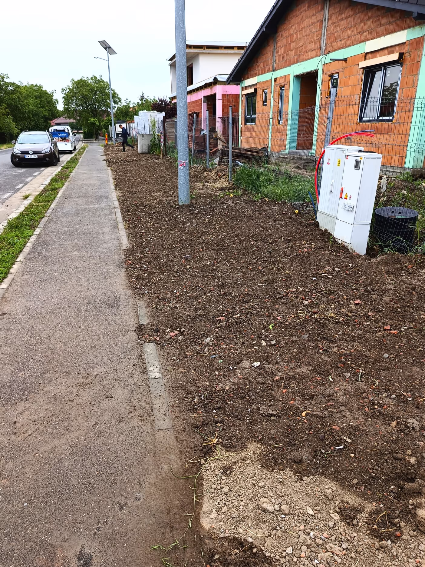A newly developed residential street with houses under construction, showing exposed brick walls, utility boxes, and a sidewalk with freshly dug soil.