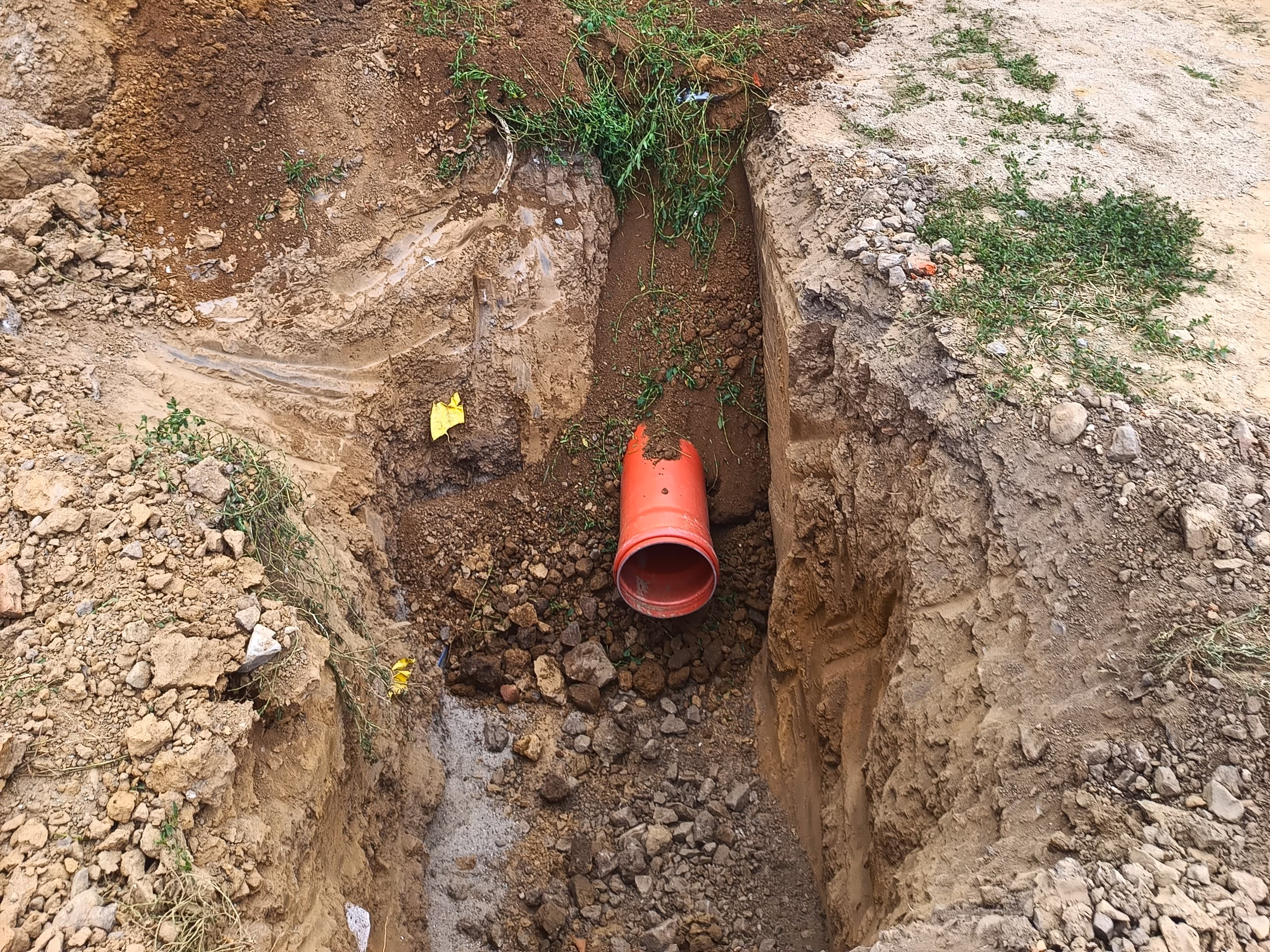 A deep trench with an orange drainage pipe partially installed, surrounded by uneven soil and construction debris.
